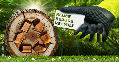 Manual worker with work gloves holding a business card with text Reuse, Reduce and Recycle. Green meadow with a cross section of a tree trunk with a recycle symbol and a green forest on background.