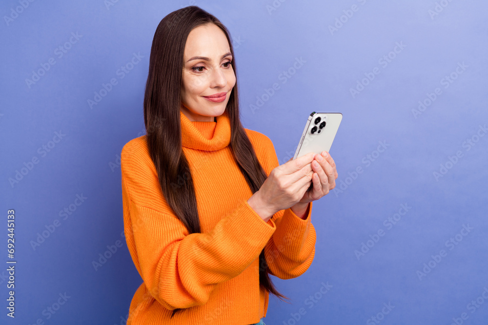 Photo of attractive young happy woman in orange pullover browsing cellphone check spam sms in gmail isolated on violet color background Photo of attractive young happy woman in orange pullover browsing cellphone check spam sms in gmail isolated on violet color background