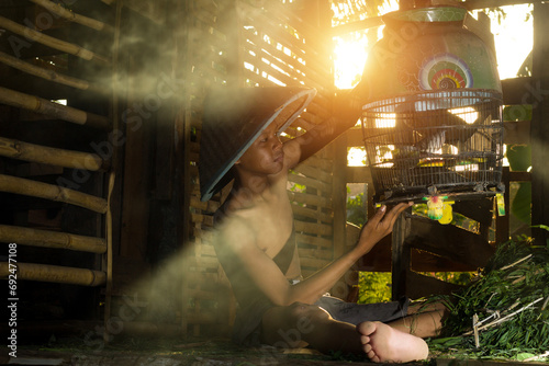 an Indonesian boy wearing a farmer's hat is repairing a bird cage with a hut in the background with afternoon lighting