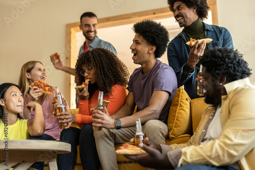 Group of multiethnic friends on a couch eating pizza and drinking beer at a house party, while laughing telling stories © PintoArt
