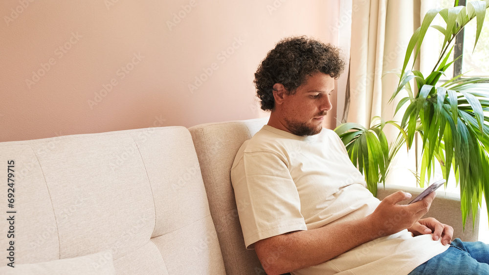 Young man with curly hair and beard, sitting on sofa in cozy living room, using mobile phone