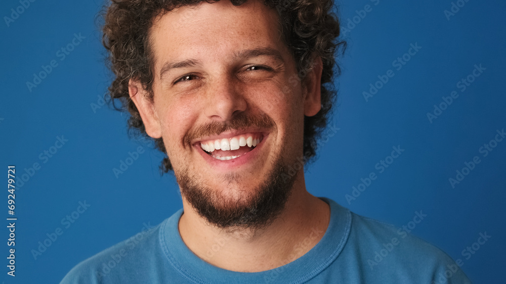 Close up, guy with curly hair dressed in blue t-shirt looks at the camera and laughs isolated on blue background in the studio Close up, guy with curly hair dressed in blue t-shirt looks at the camera and laughs isolated on blue background in the studio