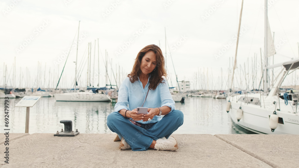 Lovely middle-aged woman is sitting in the port and using mobile phone