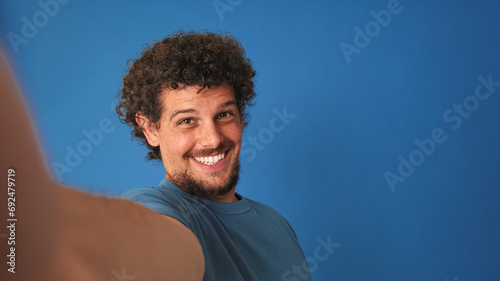 Close-up of guy with curly hair dressed in blue t-shirt, looking at the camera, making video call with mobile phone on blue background in the studio