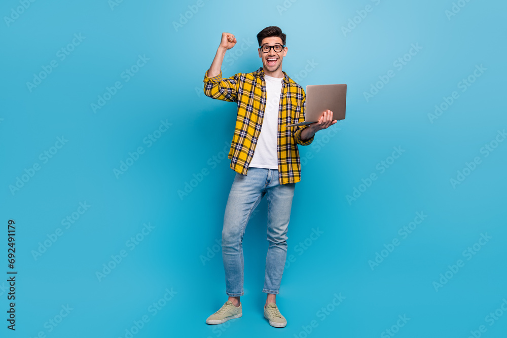 Full body photo of young businessman raised fist up enjoying playing on money betting esport hold netbook isolated on blue color background Full body photo of young businessman raised fist up enjoying playing on money betting esport hold netbook isolated on blue color background
