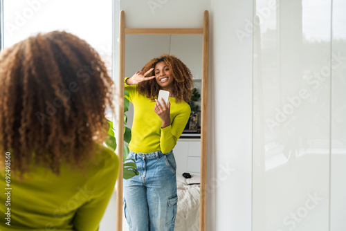 Obraz na płótnie Cheerful curly woman holding mobile phone and making selfie in the mirror w salonie