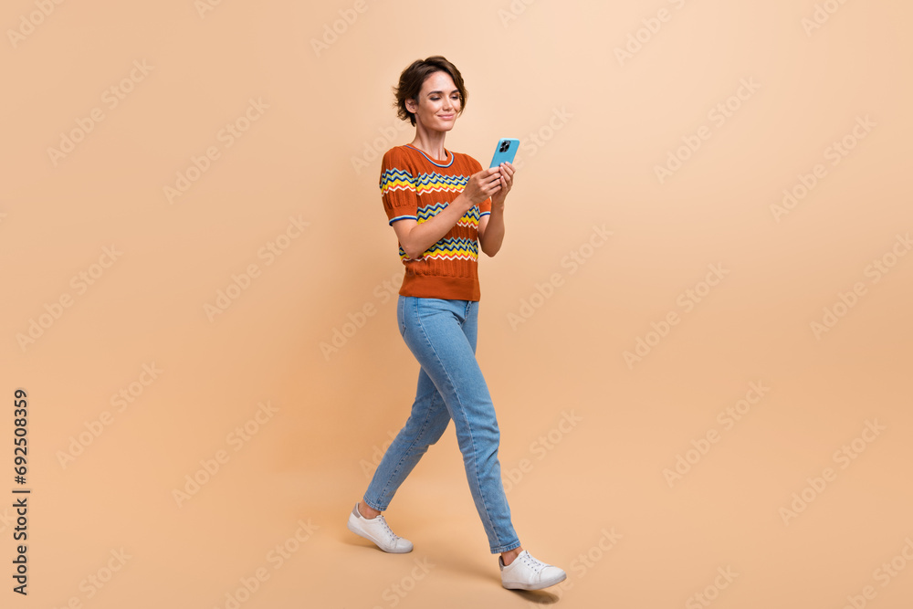 Full body profile photo of lovely positive lady walking use smart phone typing blog post isolated on beige color background Full body profile photo of lovely positive lady walking use smart phone typing blog post isolated on beige color background