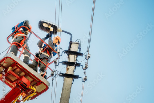 Two electricians from cradle of aerial platform or crane are repairing street lighting lamp. Professional electricians wearing helmets, overalls and insurance work at heights. View of workers from