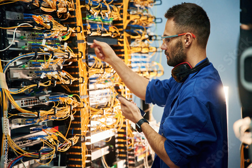 Holding smartphone. Young man is working with internet equipment and wires in server room photo