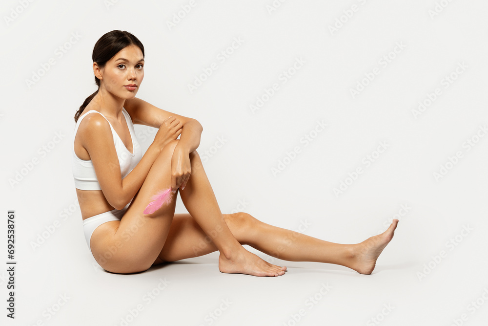 Calm asian millennial woman in white wear sits on floor, lightly brushing her leg with pink feather Calm asian millennial woman in white wear sits on floor, lightly brushing her leg with pink feather