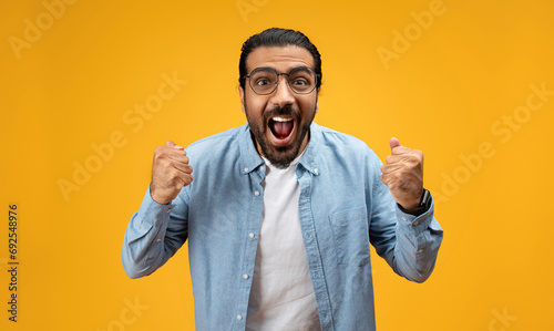 Overjoyed man in glasses and denim shirt clenches fists in victory, mouth wide open photo