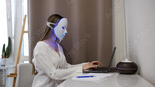 A beautiful girl with an LED mask on her head works at a laptop. Home skin care concept.