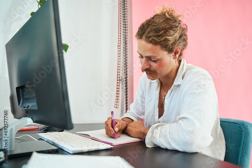 Creative young freelancer with face makeup sitting at computer table and writing notes photo