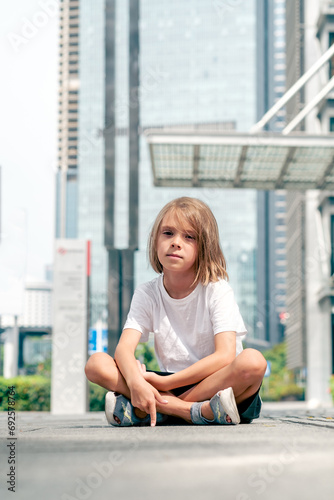 A 6-year-old girl is sitting on the asphalt in the city with an indifferent face. Growing up children in a big city