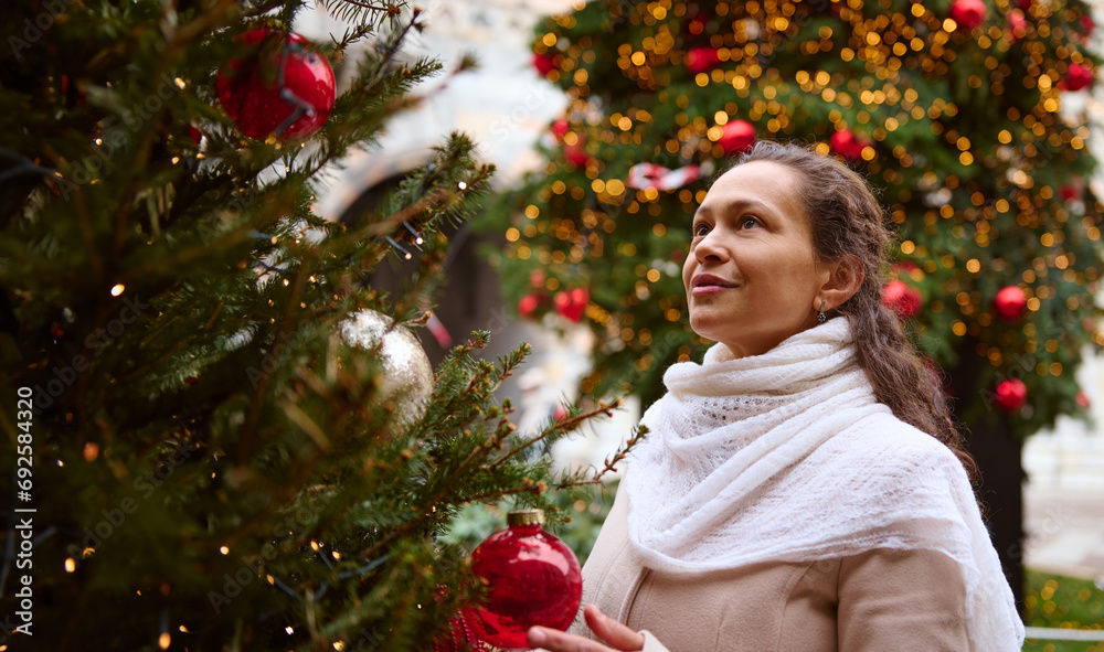 Pretty woman looking up, standing by a Christmas tree decorated with beautiful toys and garlands at Christmas funfair Pretty woman looking up, standing by a Christmas tree decorated with beautiful toys and garlands at Christmas funfair