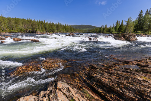 The swift Namsen River in Trondelag, Norway, flows through cascades framed by rugged shoreline stones and dense forests, all under a radiant blue sky on a sunny summer day