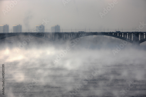 Dense frosty fog rises over the river. Majestic winter chill day in Siberia. Cityscape with road bridge with cars is visible among the fog vapors. Evaporation of water over the river at winter morning