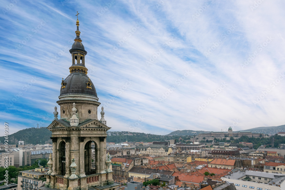 An aerial view of Budapest, featuring the corner tower of St. Stephen Basilica, rooftops of Pest, and the hills of Buda in the background