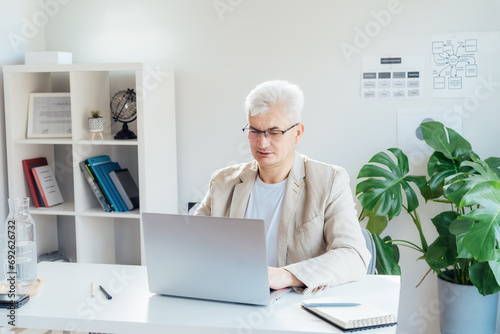 Concentrated grey hair middle-aged man working on laptop while sitting at his work place in home office. Confident, experienced senior male professional. Small business entrepreneur manage business