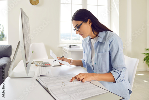 Portrait of a serious young business woman accountant in glasses sitting at the desk with pc computer with tables and charts in a light modern office on her workplace analyzing company data. photo