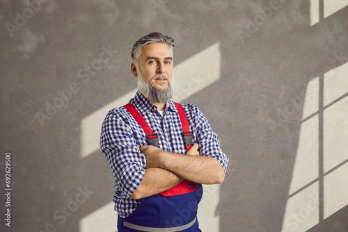 Fototapeta Naklejka Na Ścianę i Meble -  Senior repairman. Portrait of a mature male worker in uniform in the house. Bearded grey haired repair man in a shirt and blue workwear overalls standing with folded arms on a grey wall background