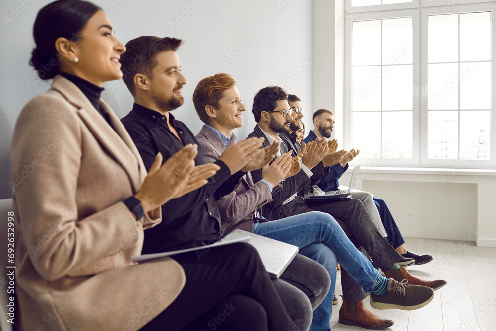 Smiling business people applauding apeaker at business conference in office. Employees sitting on chairs, clapping hands, cheering mentor, coach or team leader at corporate meeting, briefing, workshop