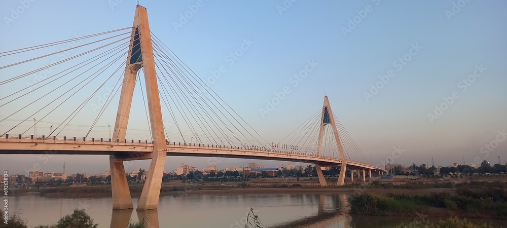 Obraz premium The view of the bridge between the river and trees and the blue sky at sunset