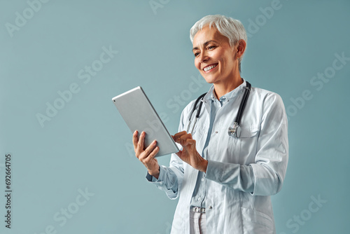 Beautiful mature female doctor in white coat holding digital tablet and looking at it, smiling while standing against blue background. Healthcare and digital technologies. photo