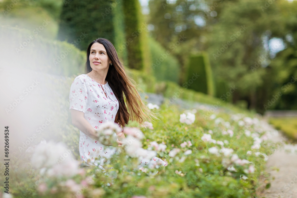 Portrait of young attractive woman sitting among white roses in picturesque old park of small town near an ancient French castle Portrait of young attractive woman sitting among white roses in picturesque old park of small town near an ancient French castle