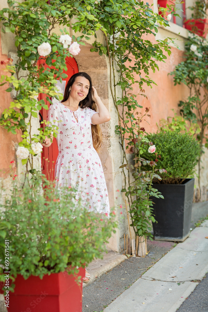 Lifestyle portrait of young stylish woman staying near red door on the street in old town and looking at white roses flowers, wearing white midi dress