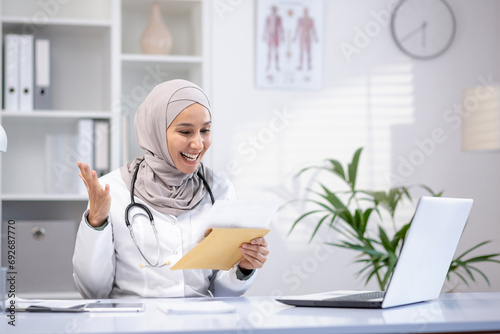 Happy female doctor in hijab holding letter in hands, sitting at table in hospital office, happy with good news. © Liubomir