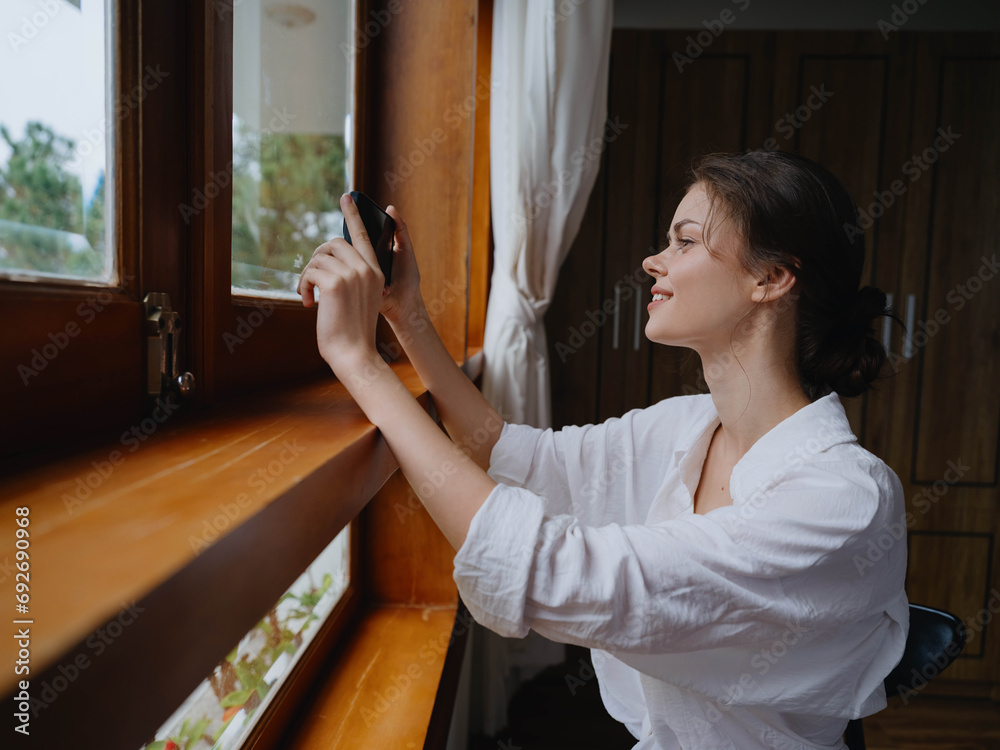 Beautiful woman with phone in hand standing at home window, freelancer on vacation, home convenient lifestyle with online work, cyber security concept, smart home. Beautiful woman with phone in hand standing at home window, freelancer on vacation, home convenient lifestyle with online work, cyber security concept, smart home.