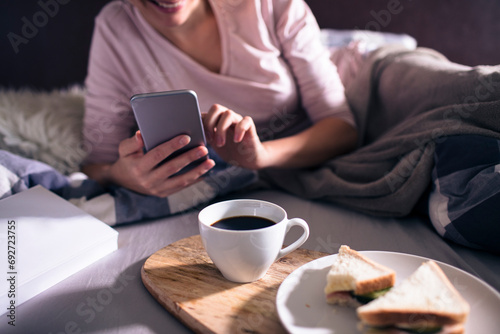 Woman having breakfast in bed using smartphone