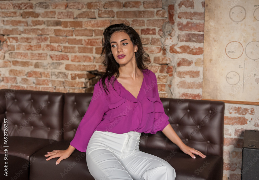 Beautiful young brunette woman in a purple blouse and white pants posing indoor against a brick wall. Portrait of a beautiful young brunette woman