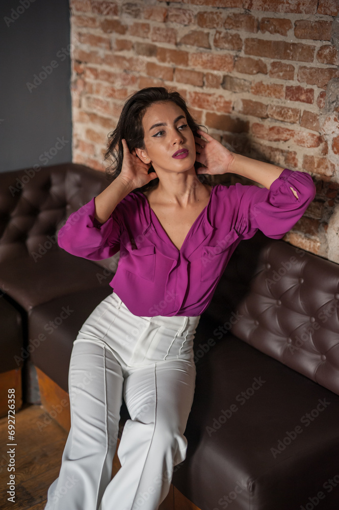 Beautiful young brunette woman in a purple blouse and white pants posing indoor against a brick wall. Portrait of a beautiful young brunette woman