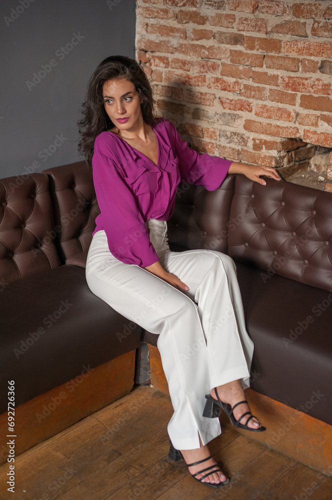 Beautiful young brunette woman in a purple blouse and white pants posing indoor against a brick wall. Portrait of a beautiful young brunette woman