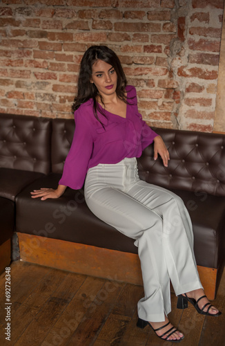 Obraz na płótnie Beautiful young brunette woman in a purple blouse and white pants posing indoor against a brick wall. Portrait of a beautiful young brunette woman w salonie