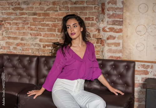 Obraz na płótnie Beautiful young brunette woman in a purple blouse and white pants posing indoor against a brick wall. Portrait of a beautiful young brunette woman w salonie