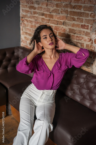 Beautiful young brunette woman in a purple blouse and white pants posing indoor against a brick wall. Portrait of a beautiful young brunette woman 