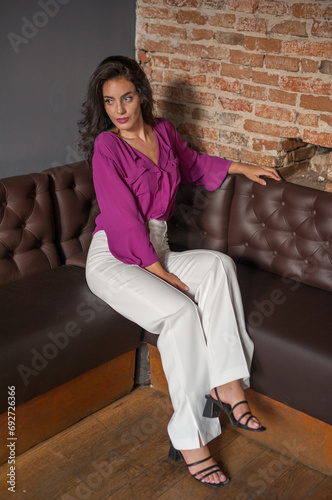 Obraz na płótnie Beautiful young brunette woman in a purple blouse and white pants posing indoor against a brick wall. Portrait of a beautiful young brunette woman w salonie