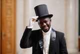 Waist up portrait of young Black gentleman wearing top hat smiling at camera in palace and giving well mannered bow