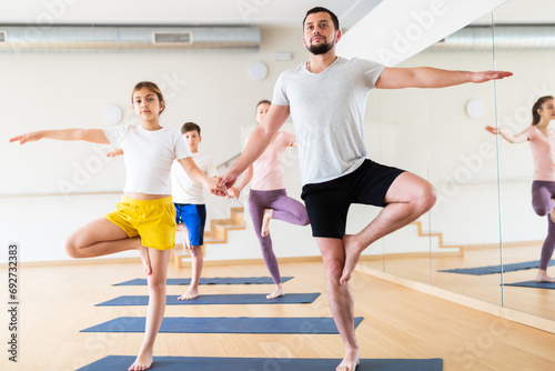 Friendly sporty family of four practicing partner yoga in fitness studio. Teen girl with father standing on one leg in Eka Pada Pranamasana pose holding hands