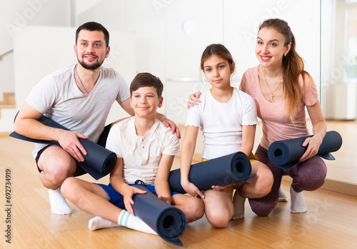 Portrait of happy sporty family with two preteen children posing in gym with yoga mats in hands..