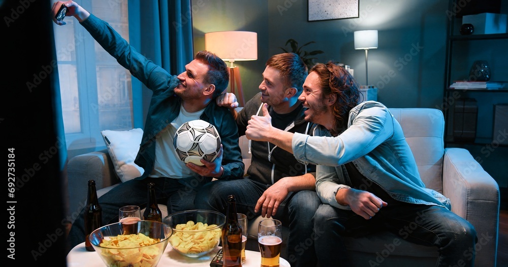 Fototapeta premium Three cheerful Caucasian friendly men sitting on sofa at night with football ball, snacks and beer and taking selfie photo with smartphone camera. Sport fans posing to phone in living room at TV.