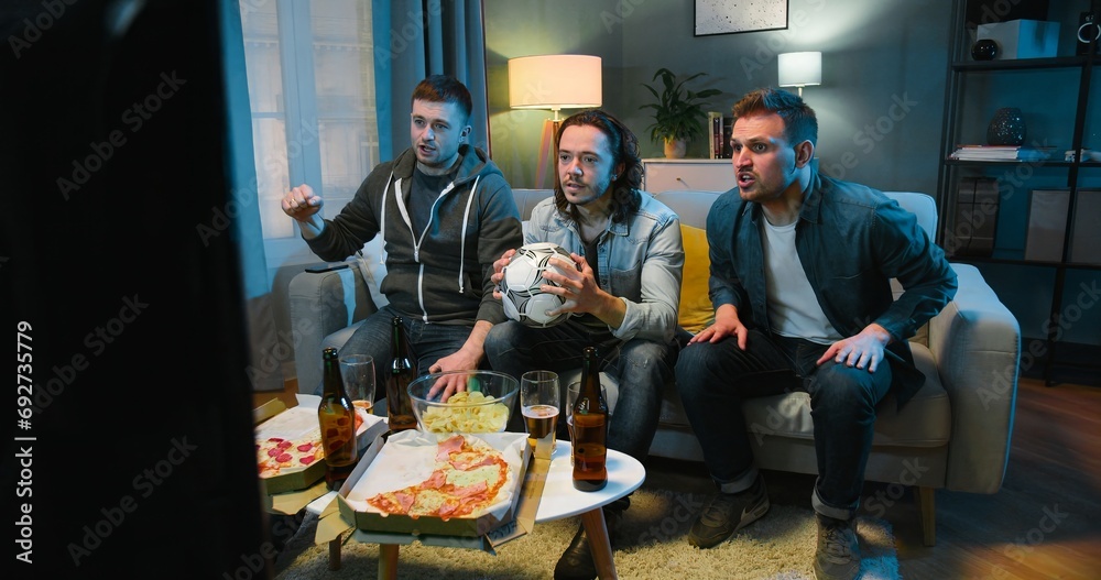 Obraz premium Caucasian joyful male friends eating pizza and doing cheers gesture with glasses of beer. Guys sitting on sofa in living room at night in front of TV. Football fans watching match on sport channel.