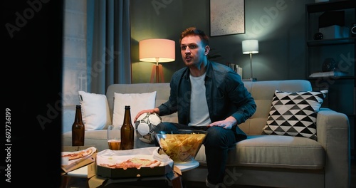 Caucasian young guy sitting on sofa at night in dark living room, eating potato chips and drinking beer. Male football fan is angry as favorite team loosing or missing goal. Man watching TV
