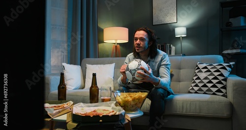 Happy cheerful Caucasian man sport fan sitting on couch in evening at home and watching football match on TV. Guy eating potato chips and drinking beer. Favorite team winning or scoring goal concept.