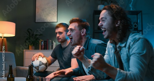 Tensed Caucasian male football fans screaming, worrying and cheering for favorite team while watching TV and sitting late at night in living room. Guys friends having nice evening together at home. photo