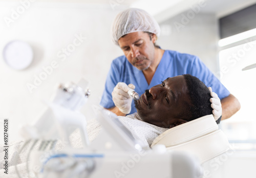 African man receiving facial cleansing procedure with attachment producing water with hydrogen and oxygen ions, ensuring deep penetration into dermis. Modern hardware cosmetology
