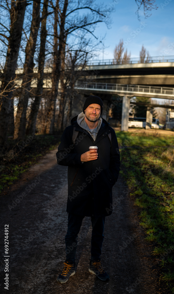 Caucasian male in a coat holding coffee standing on a park trail Caucasian male in a coat holding coffee standing on a park trail